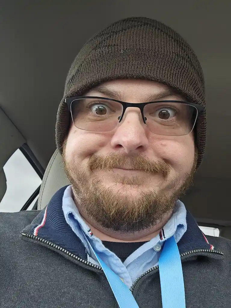 A man wearing a brown beanie, glasses, and a beard sits in a car, making a wide-eyed, exaggeratedly shocked face while…