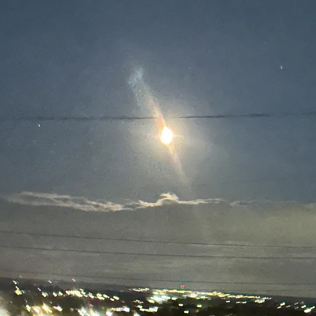 A blurry photo of the sun or moon shining through a hazy sky with power lines and distant city lights below. The image has a…