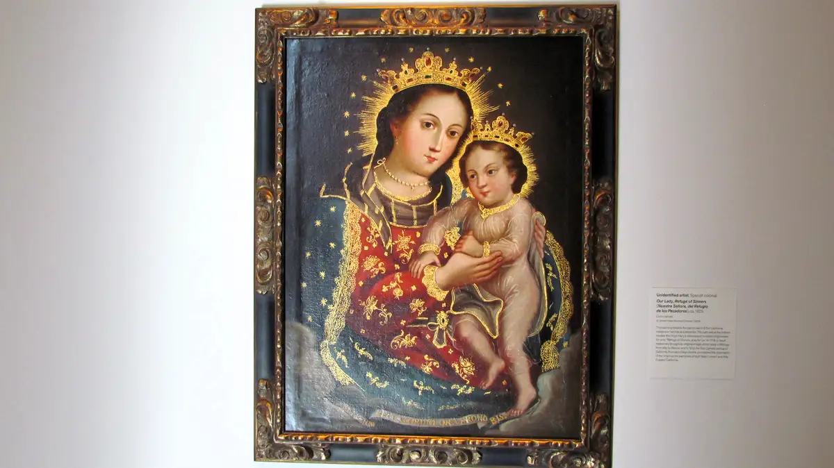 A framed religious painting depicts the Virgin Mary holding the infant Jesus, both wearing crowns and halos, set against a…