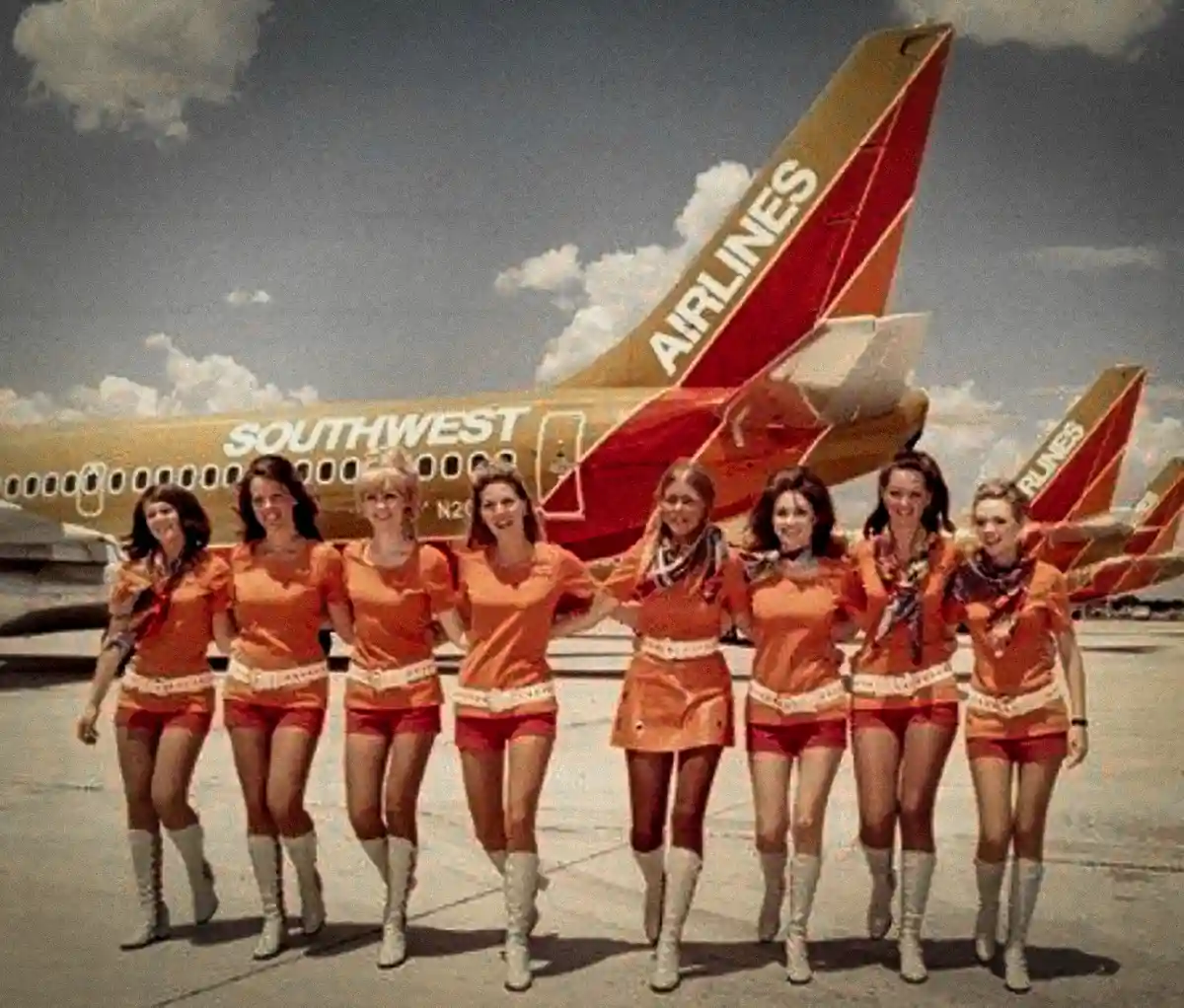 A vintage photograph shows seven female flight attendants in bright orange uniforms with white belts and tall white boots…