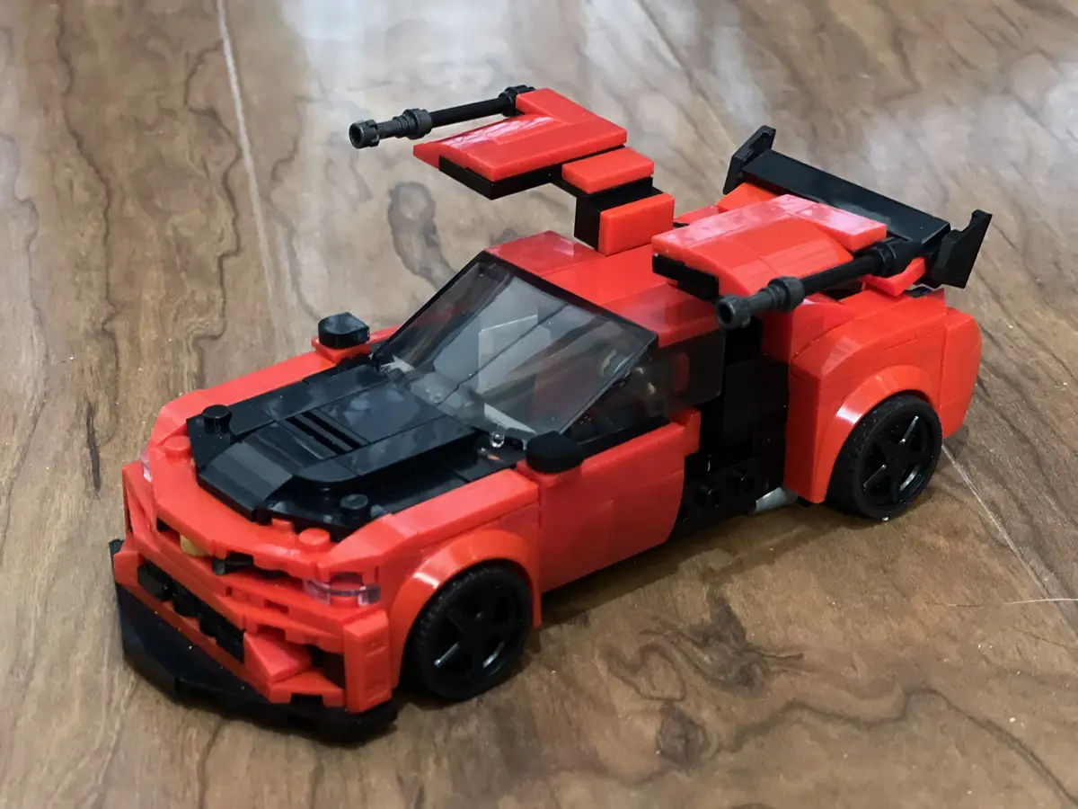 A red and black Lego model of a sports car sits on a wooden surface, featuring two black toy guns mounted on top of its…