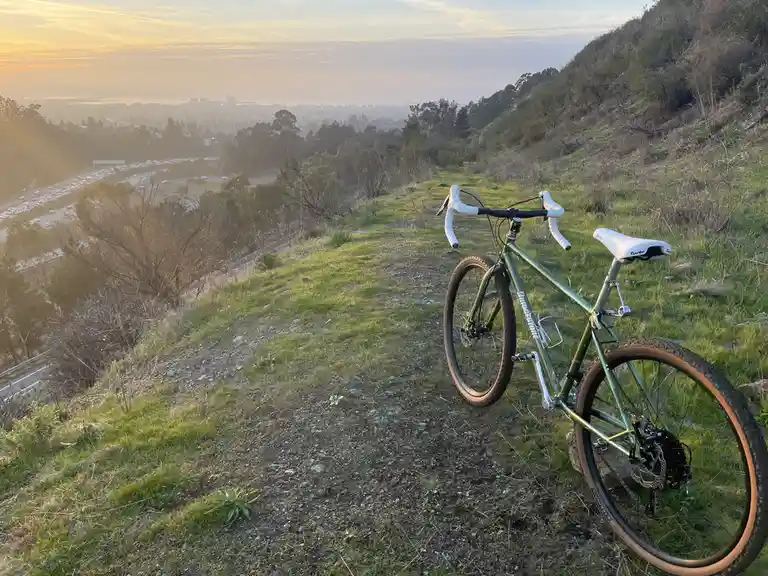 A green bicycle with white handlebar tape and saddle is parked on a grassy hillside overlooking a busy highway filled with…