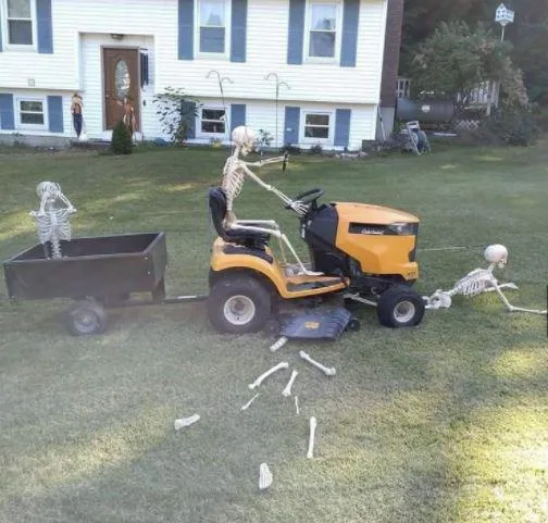 Three plastic skeleton decorations are arranged on a grassy lawn in front of a house. One skeleton sits in the driver's seat…