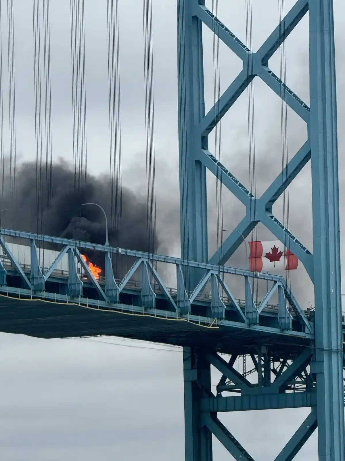 A photo of the Ambassador Bridge shows a large fire with black smoke billowing from the roadway, while a Canadian flag is…