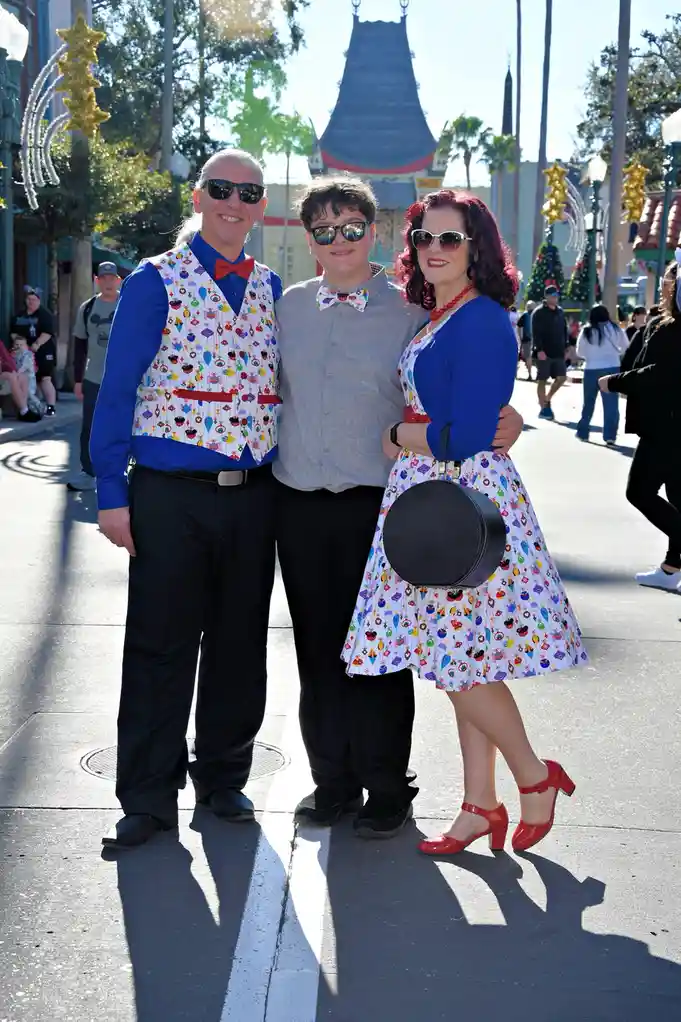 Three people pose together outdoors in a theme park, all wearing matching outfits with a colorful pattern of cartoonish hot…