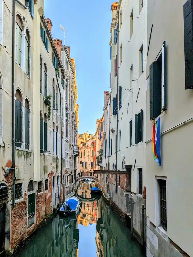 A picturesque view down a narrow Venetian canal flanked by tall, aged buildings with arched windows and green shutters. A…