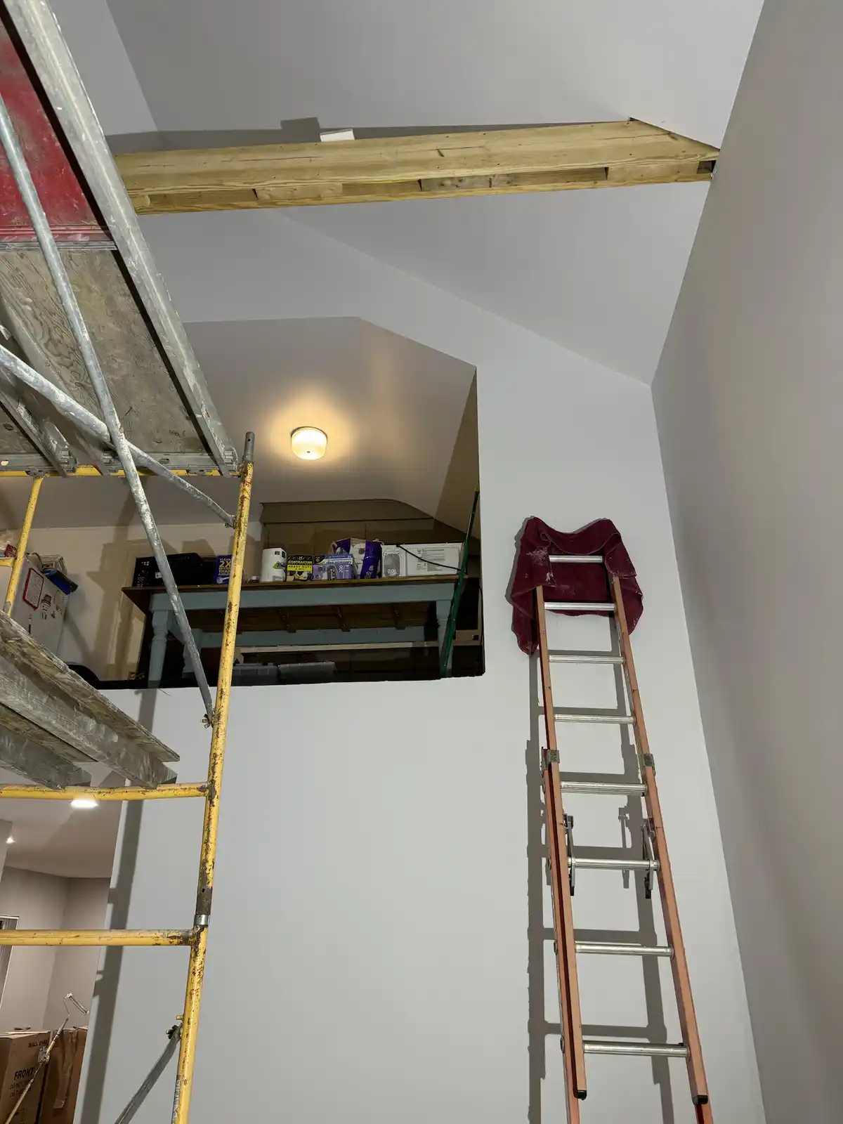 A room under renovation with a yellow scaffold on the left and a tall wooden ladder on the right, both leaning against white…
