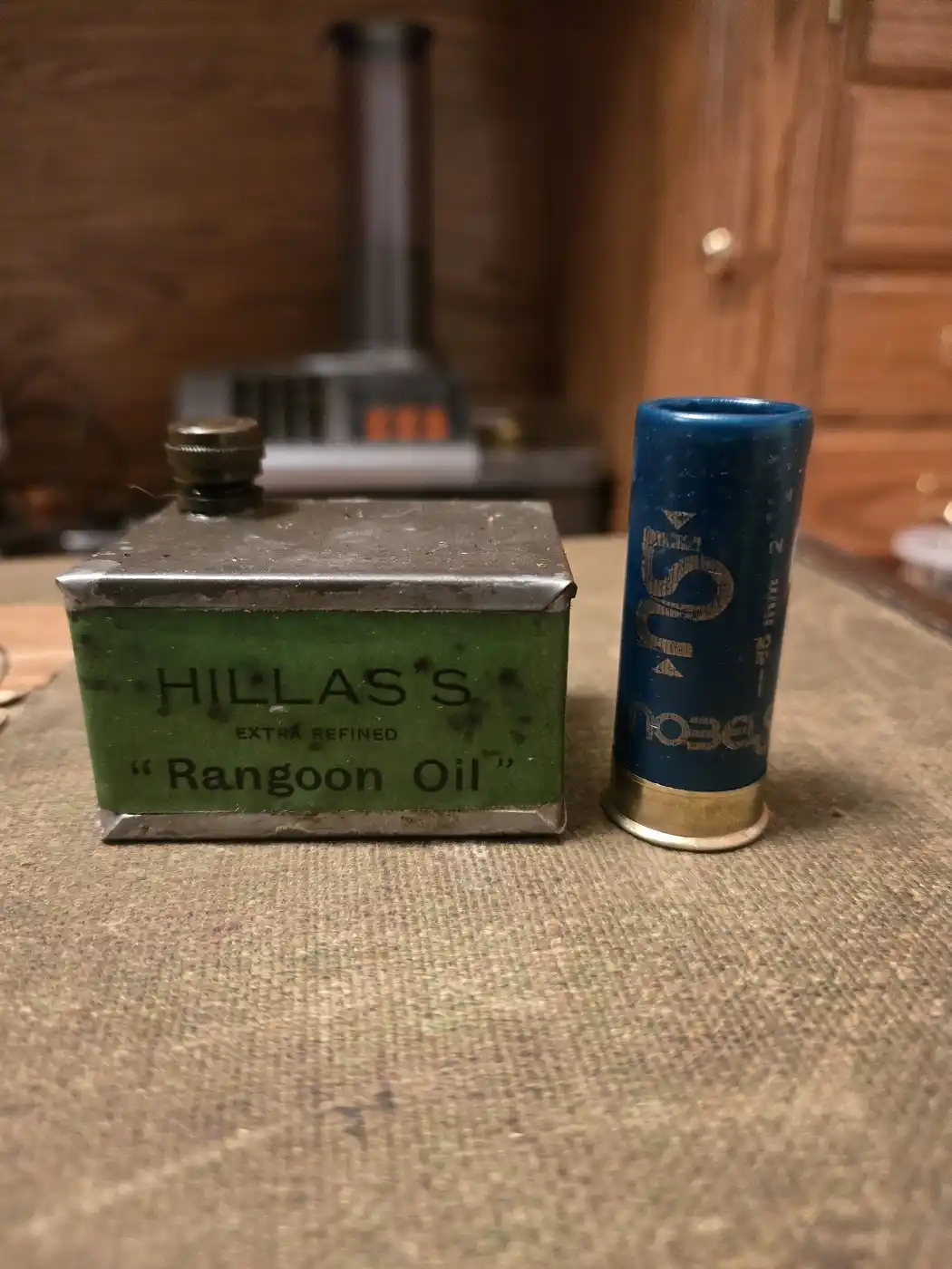 A vintage green metal tin labeled 'HILLAS'S EXTRA REFINED "Rangoon Oil"' sits next to a blue shotgun shell on a textured…