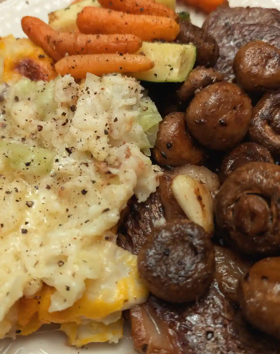 A close-up photo of a plated meal featuring mashed potatoes with cheese and green vegetables, baby carrots, zucchini…