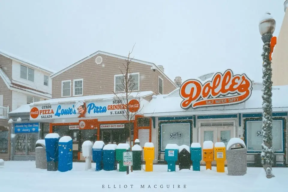 A snowy street scene shows a row of colorful, snow-capped parking meters in front of two adjacent storefronts. The left…