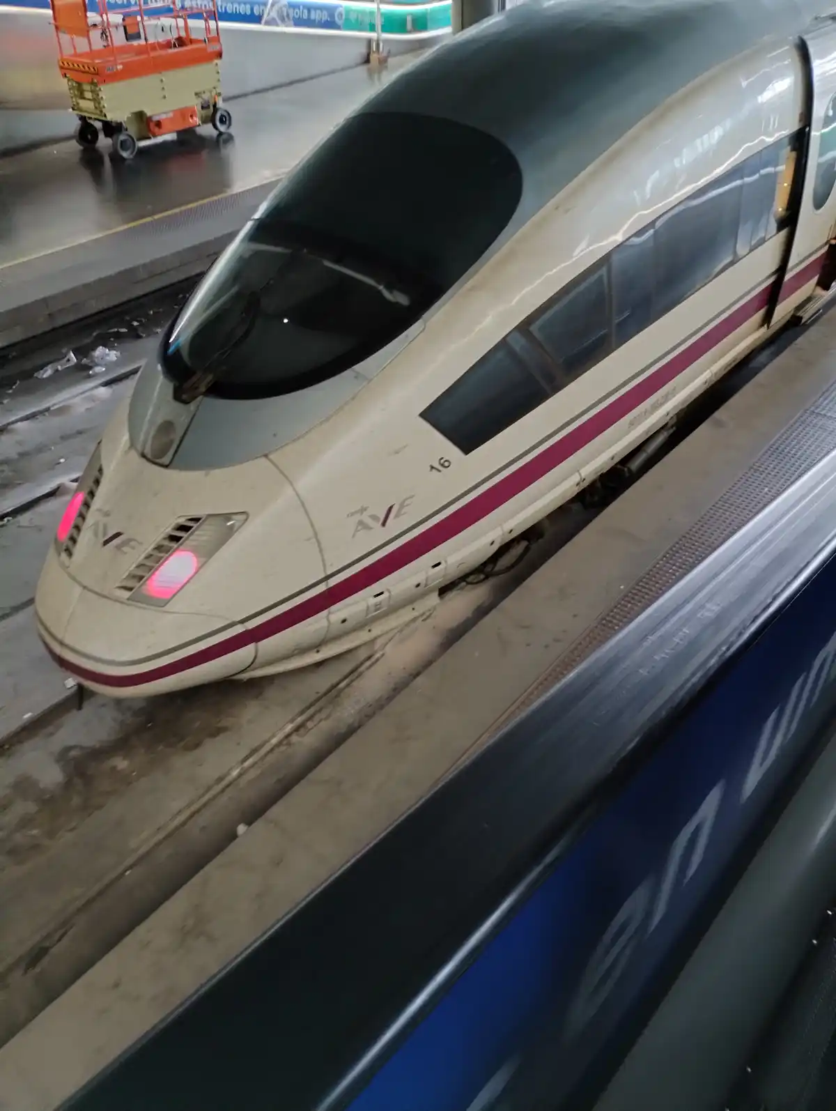 A high-speed AVE train is stationary at a station platform, its front facing the viewer. The train's headlights are on, and…