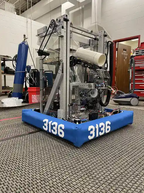 A large, complex robot with a blue padded base marked with the number 3136 sits in a workshop. The robot has a metal frame…