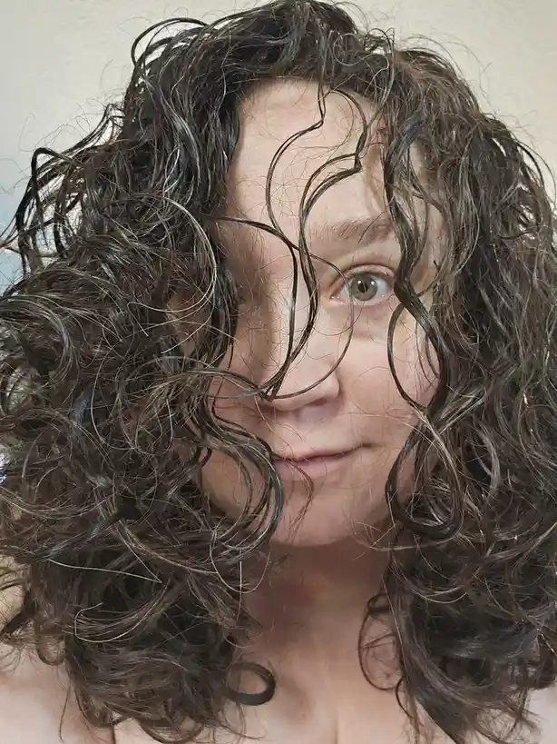 A person with dark, curly, wet-looking hair has strands covering most of their face, leaving only one eye and part of their…