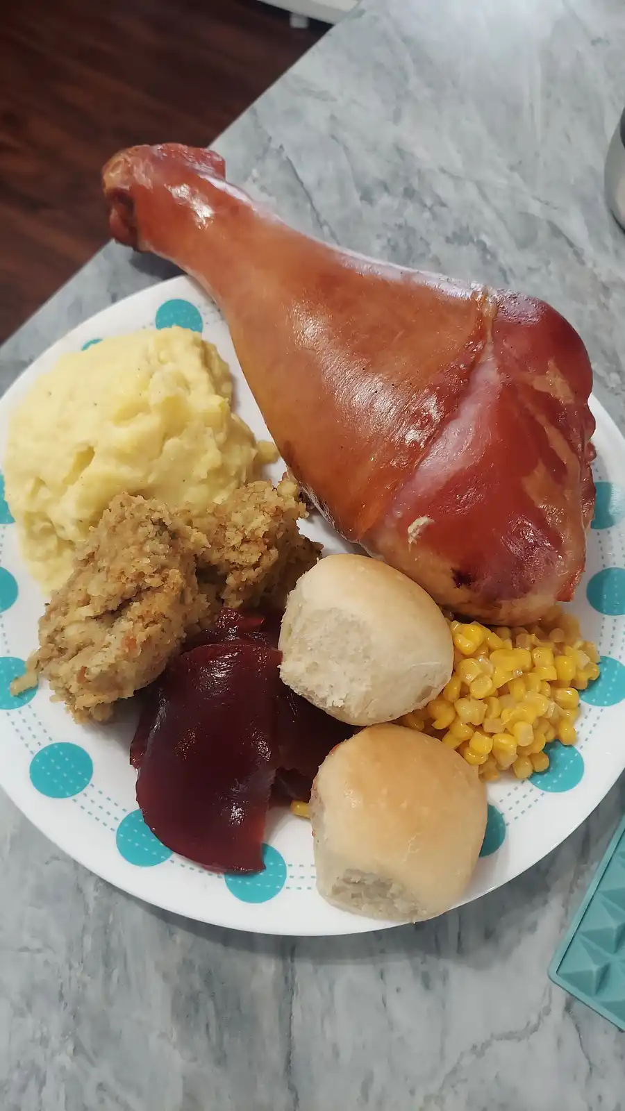 A plate of food featuring a large roasted ham leg, mashed potatoes, corn, two dinner rolls, cranberry sauce, and stuffing…