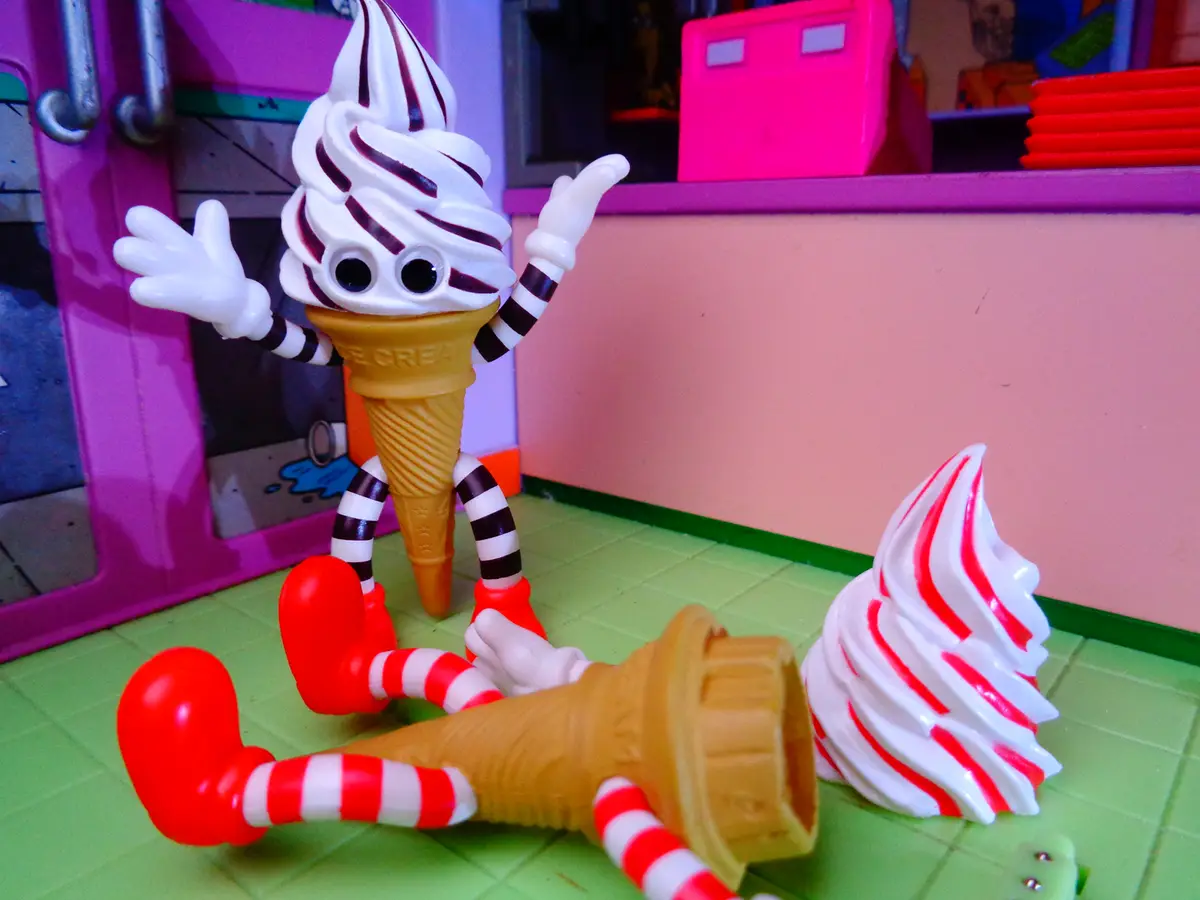 A toy ice cream cone character with black-and-white striped arms and red shoes stands upright with arms raised, while…