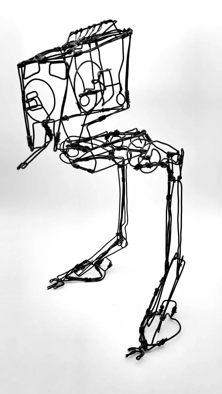 A black wire sculpture of a human figure in a hunched, slumped posture, with a box-like head, set against a plain white…
