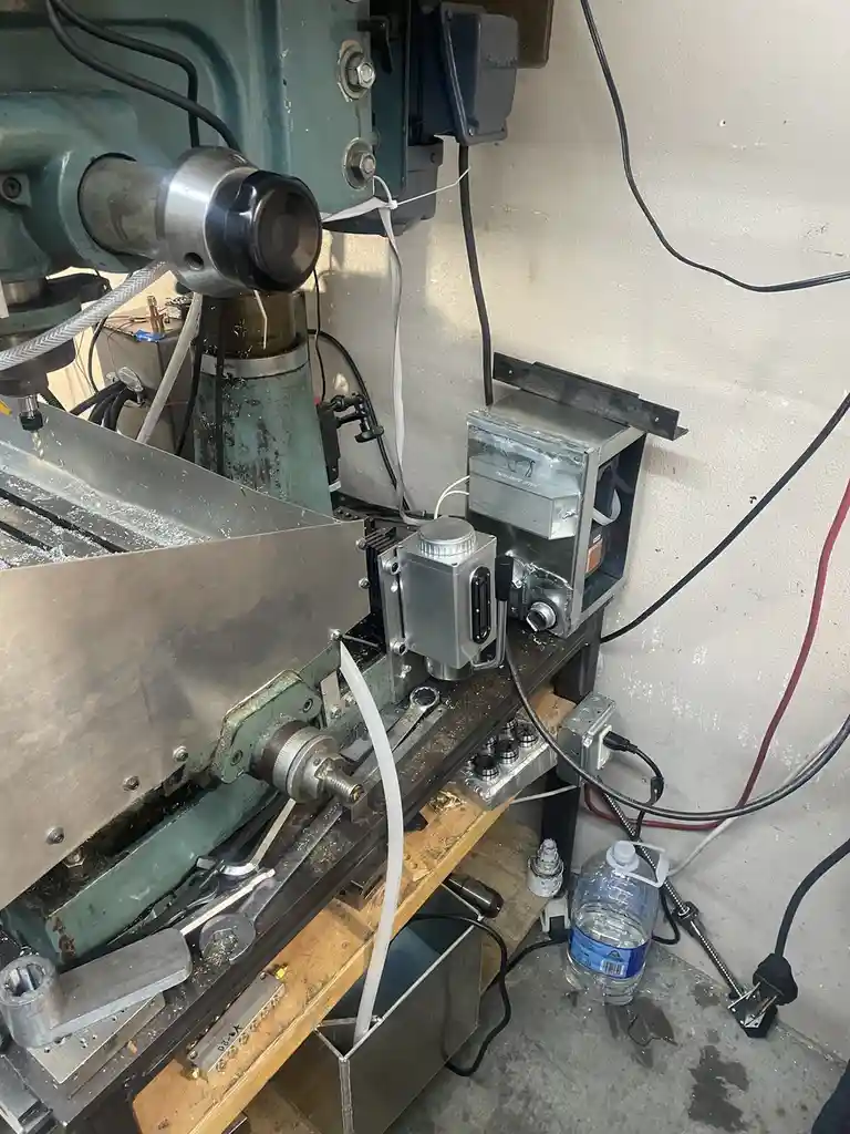 A close-up photo of a cluttered workshop showing a large, green metal milling machine with a metal workpiece clamped to its…