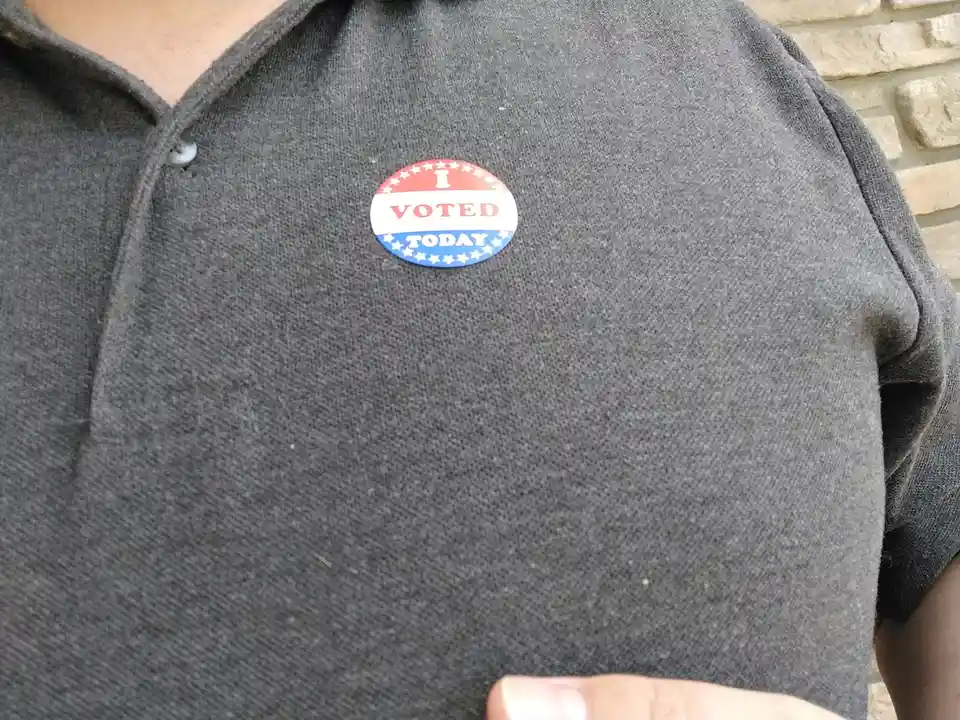 A close-up photo shows a person wearing a dark gray polo shirt with a red, white, and blue oval sticker pinned to the chest…