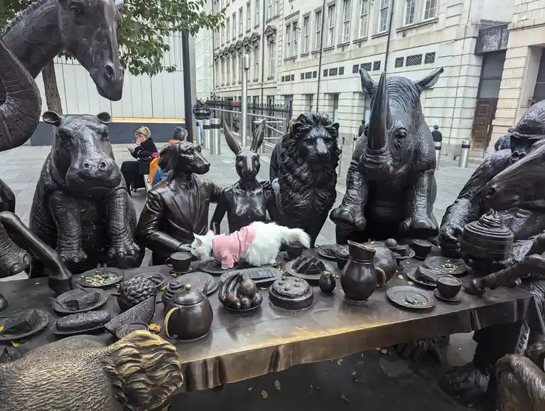 A real white cat wearing a pink sweater is sitting at a bronze statue of animals having tea, with text overlaid that reads…