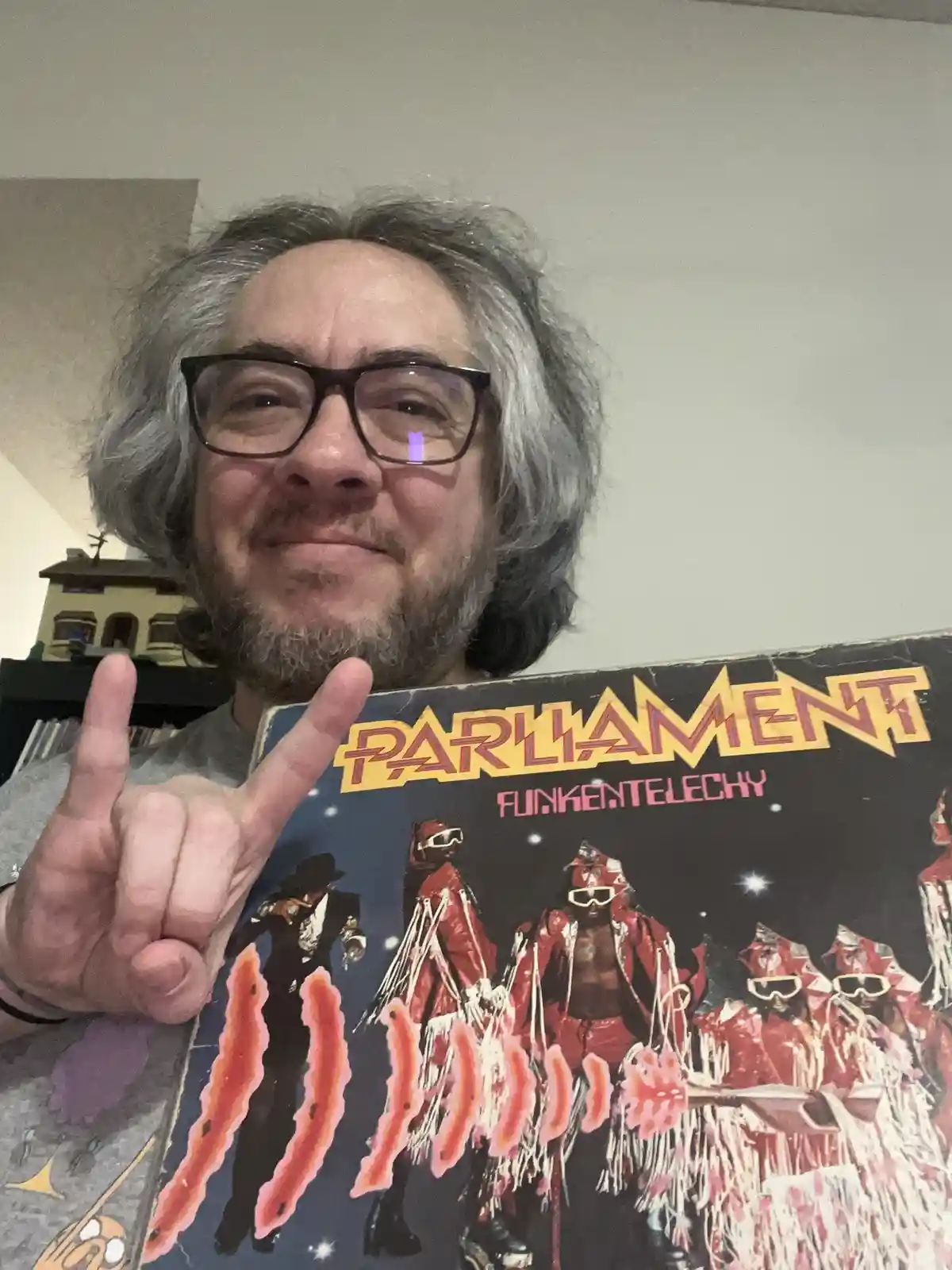 A man with gray hair and glasses holds up a 'Parliament - Funkentelechy' album cover, making a rock hand gesture. The album…