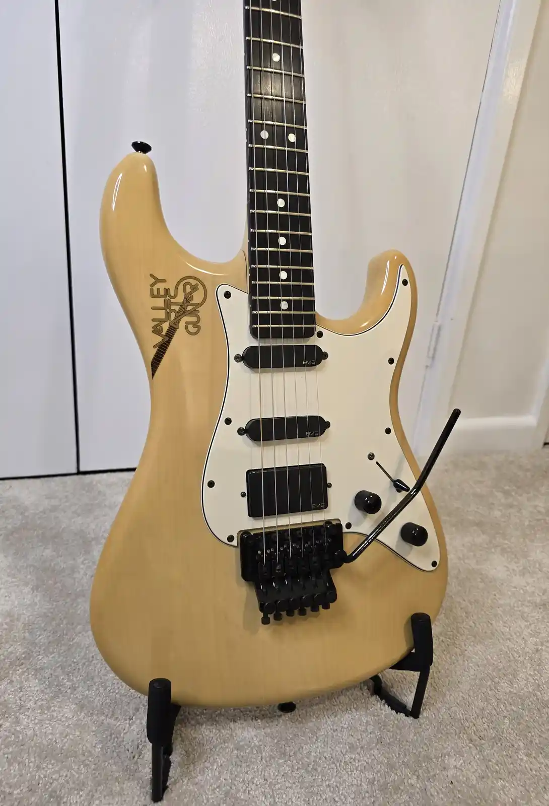 A light-wood electric guitar with a white pickguard and black hardware stands upright on a black stand against a white wall…