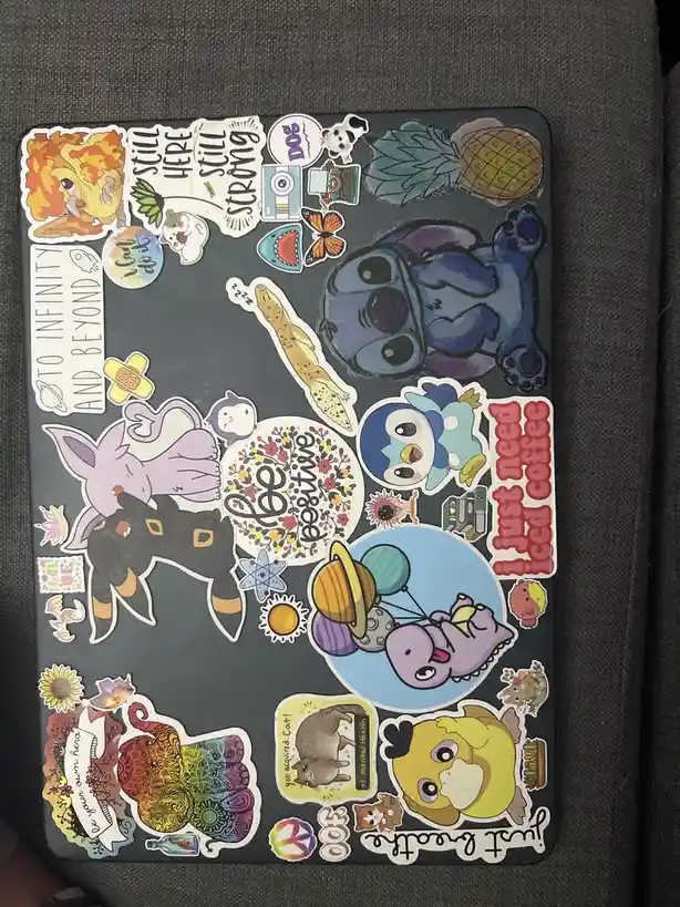 A dark gray laptop cover is densely covered with colorful stickers featuring cartoon characters like Stitch, Piplup, and…