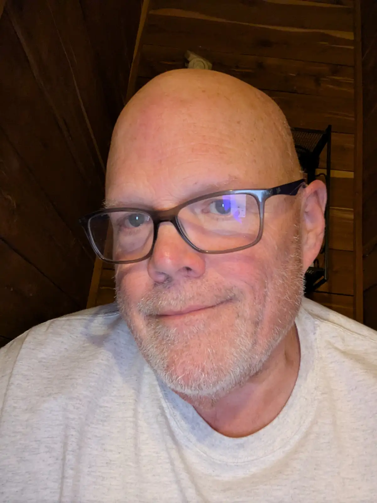 A close-up selfie of a bald man with glasses and a gray beard, wearing a light gray t-shirt, smiling slightly. The…