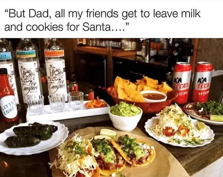 A bar counter is laden with Mexican food including tacos, guacamole, chips, and drinks like tequila, Tecate beer, and hot…