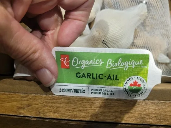 A close-up photo shows a hand holding a green and white product label for 'Organics Biologique GARLIC • AIL'. The label…