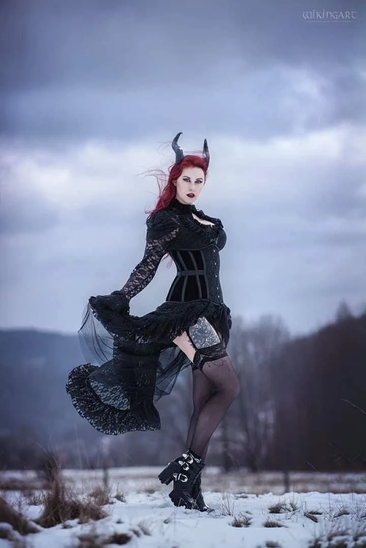 A woman with red hair and black horns stands in a snowy field wearing a black lace corset dress, thigh-high stockings, and…