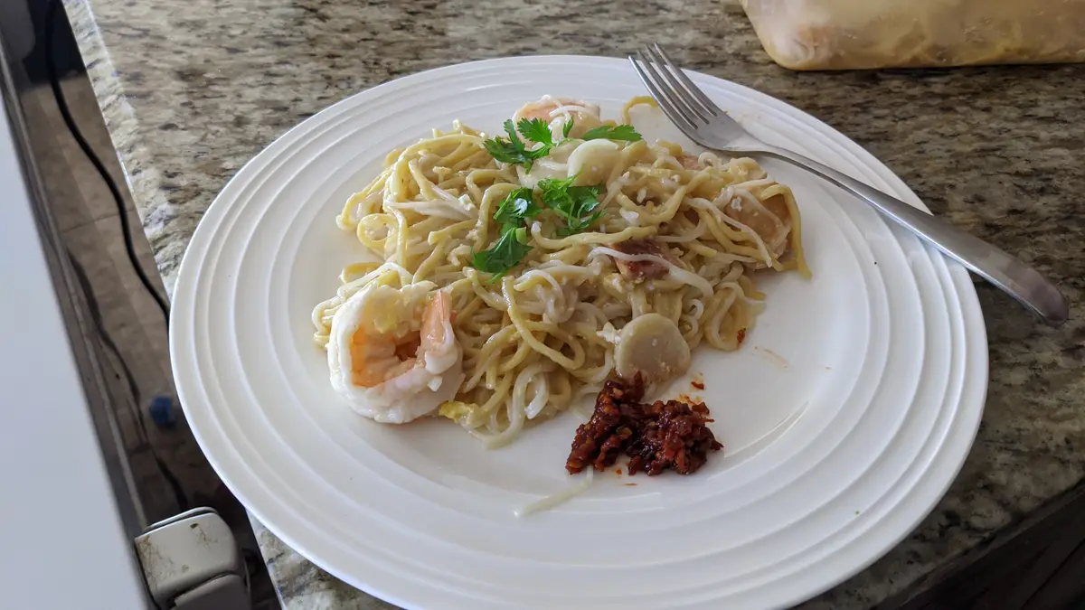 A white plate holds a serving of pasta with shrimp, scallops, and parsley, with a small pile of red chili paste on the side…
