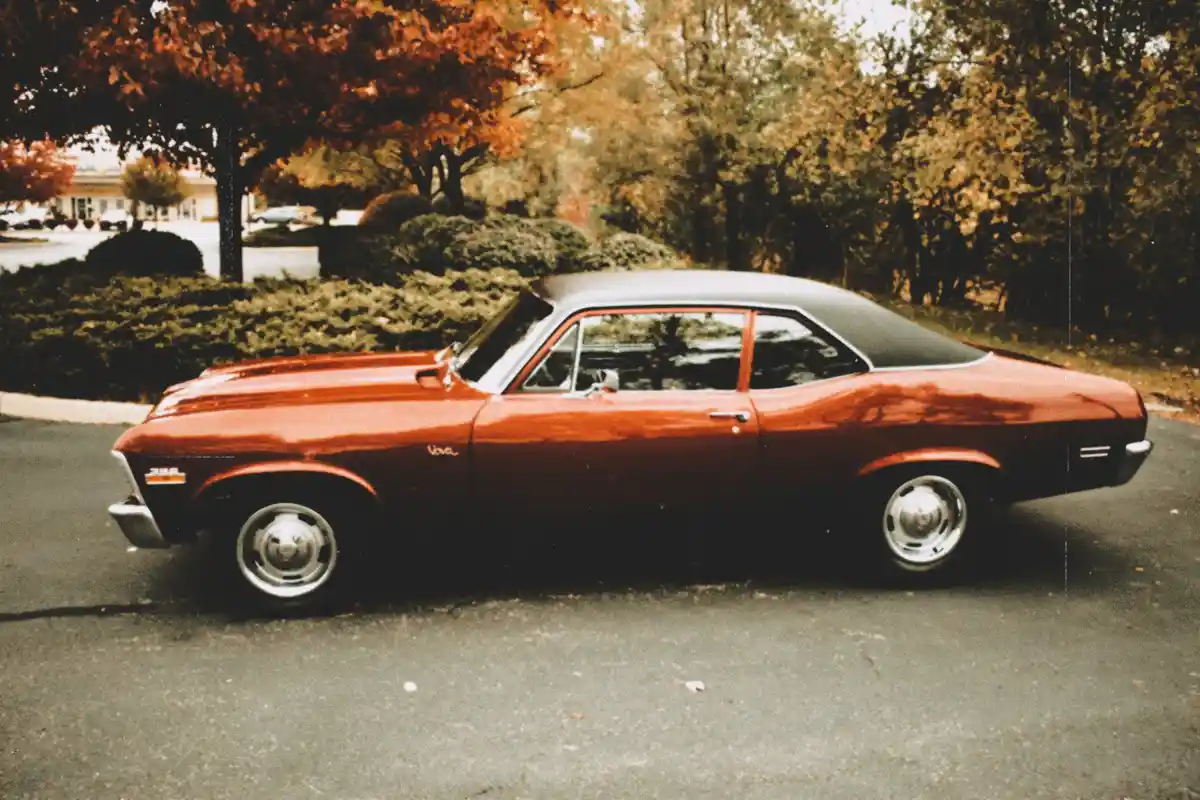 A vintage red Chevrolet Nova coupe with a black vinyl roof is parked on an asphalt surface, surrounded by trees with autumn…