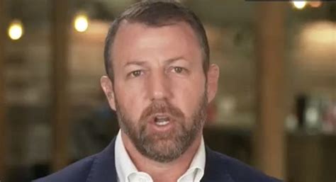 A man with a beard and short hair, wearing a dark suit jacket over a white collared shirt, is shown mid-sentence with a…