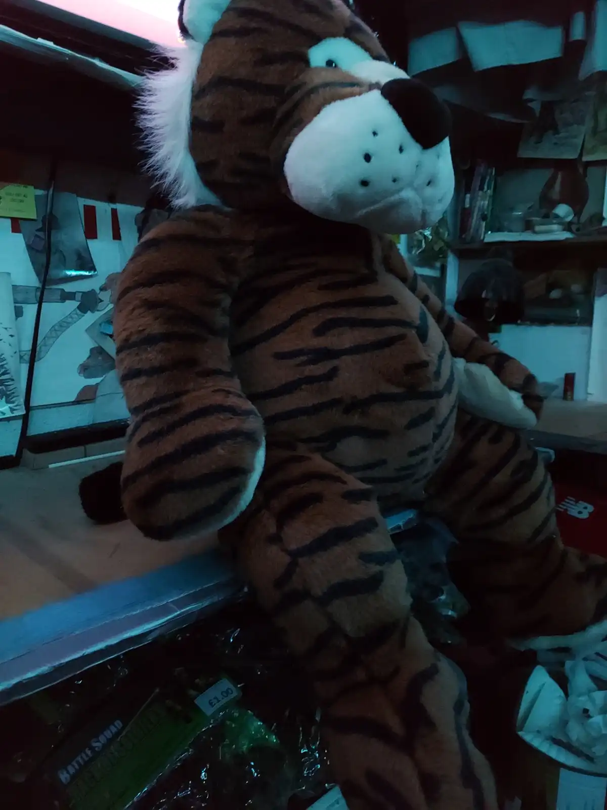 A large, brown and black striped plush tiger sits on a shelf in a cluttered store, looking slightly sad or confused. The…