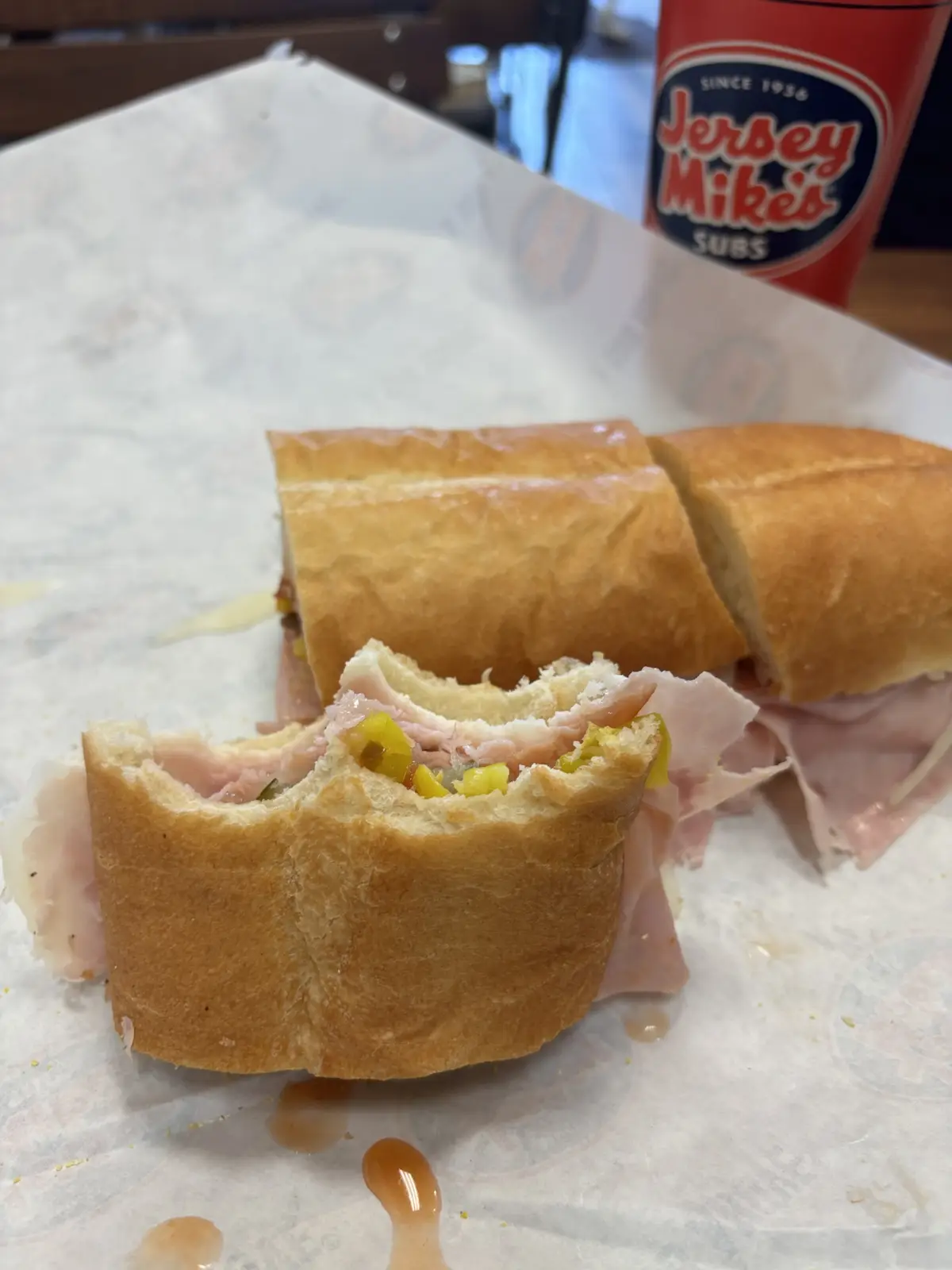 A partially eaten Jersey Mike's sub sandwich sits on branded paper, with a bite taken out of the front piece revealing ham…