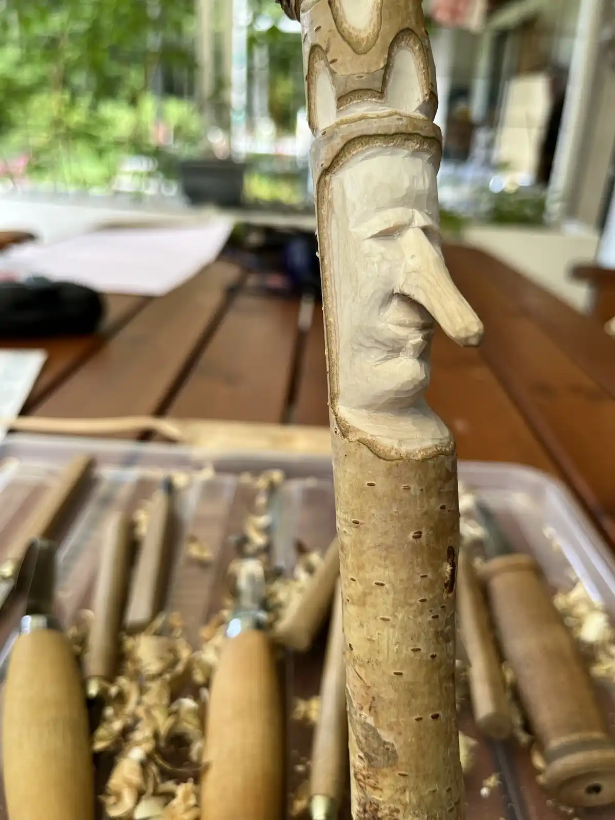 A close-up photo shows a partially carved wooden stick with a crude, unfinished human face, featuring a long nose and simple…