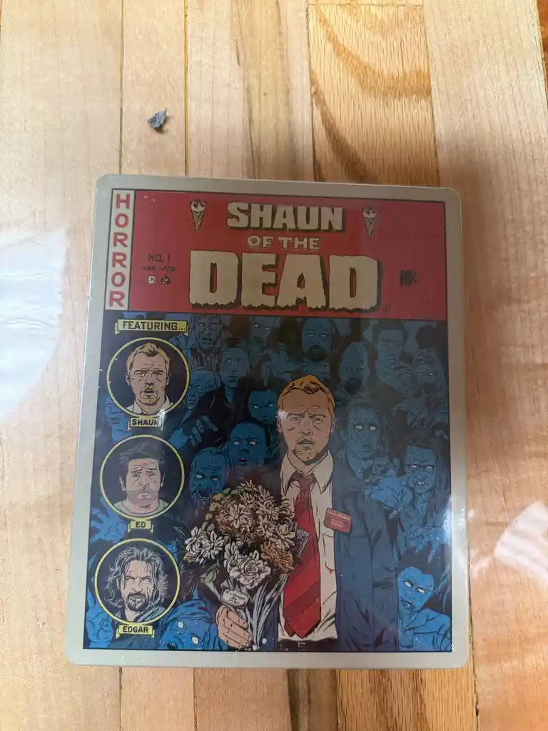 A metal tin featuring a comic book cover for 'Shaun of the Dead,' showing a man in a suit holding flowers surrounded by…