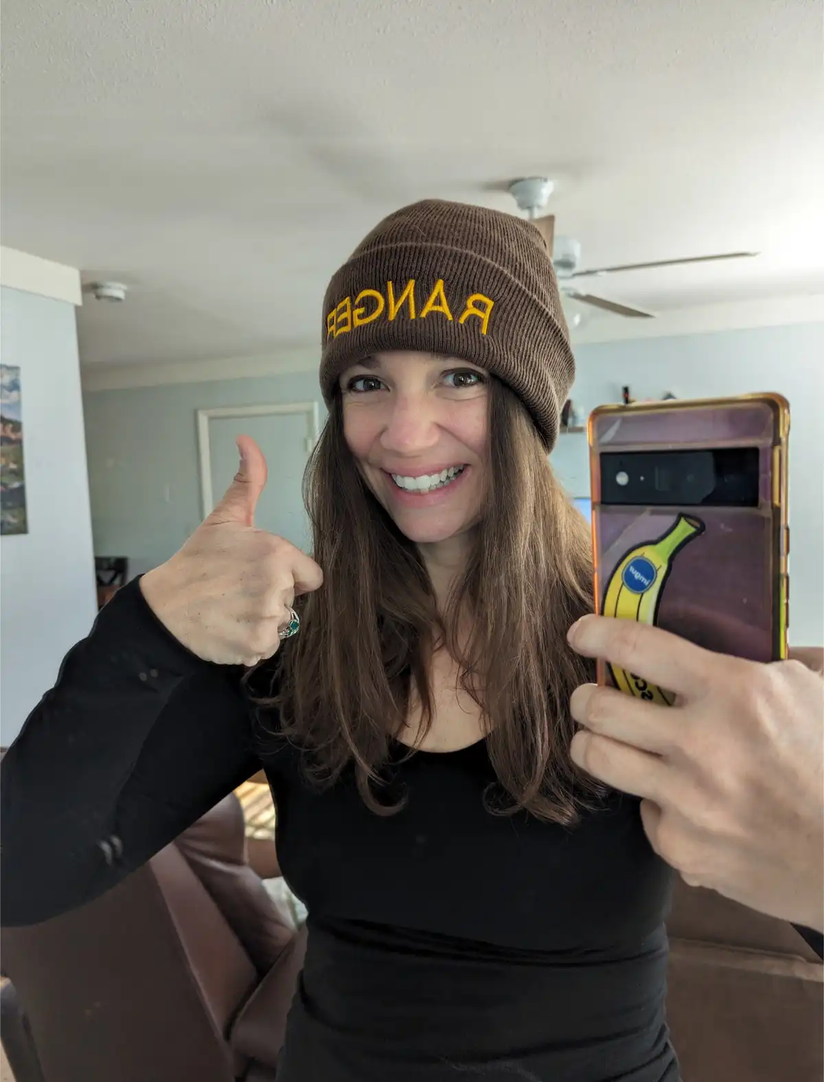 A woman with long brown hair wearing a brown beanie with 'RANGER' written in yellow backwards smiles and gives a thumbs-up…