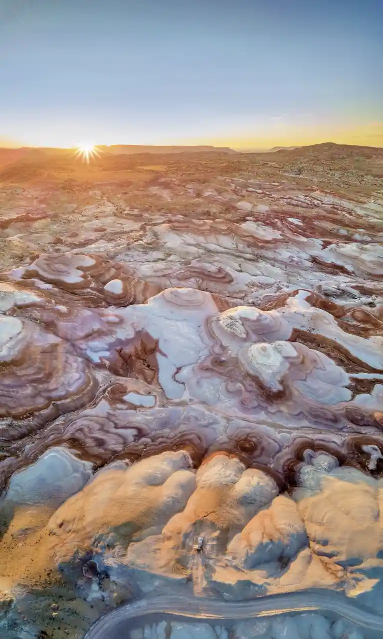 An aerial view shows a small white vehicle parked on a winding dirt road amidst vast, colorful, undulating badlands with…
