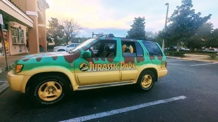 A yellow and green SUV is parked in a lot, decorated with Jurassic Park branding including the logo and camouflage pattern…