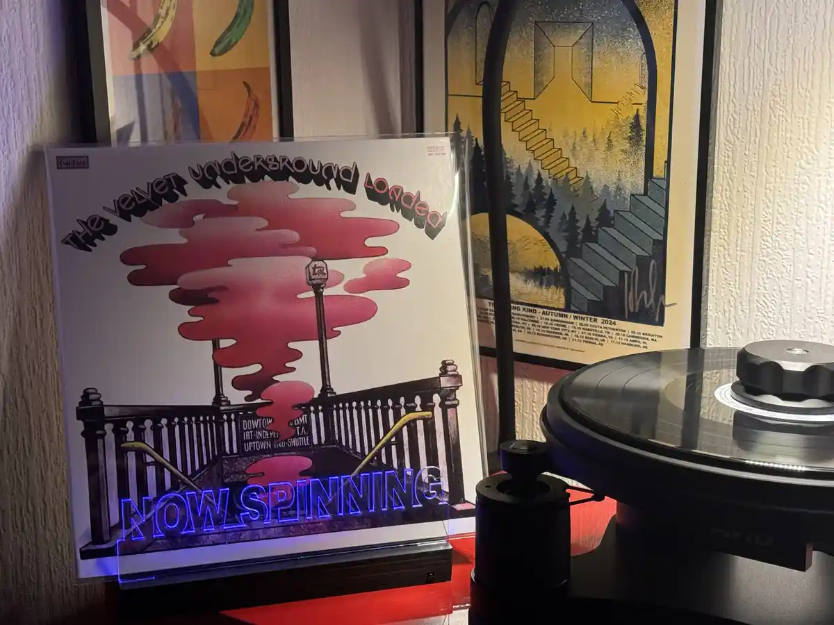 A vinyl record player with a record on the turntable sits next to a framed copy of The Velvet Underground's 'Loaded' album…