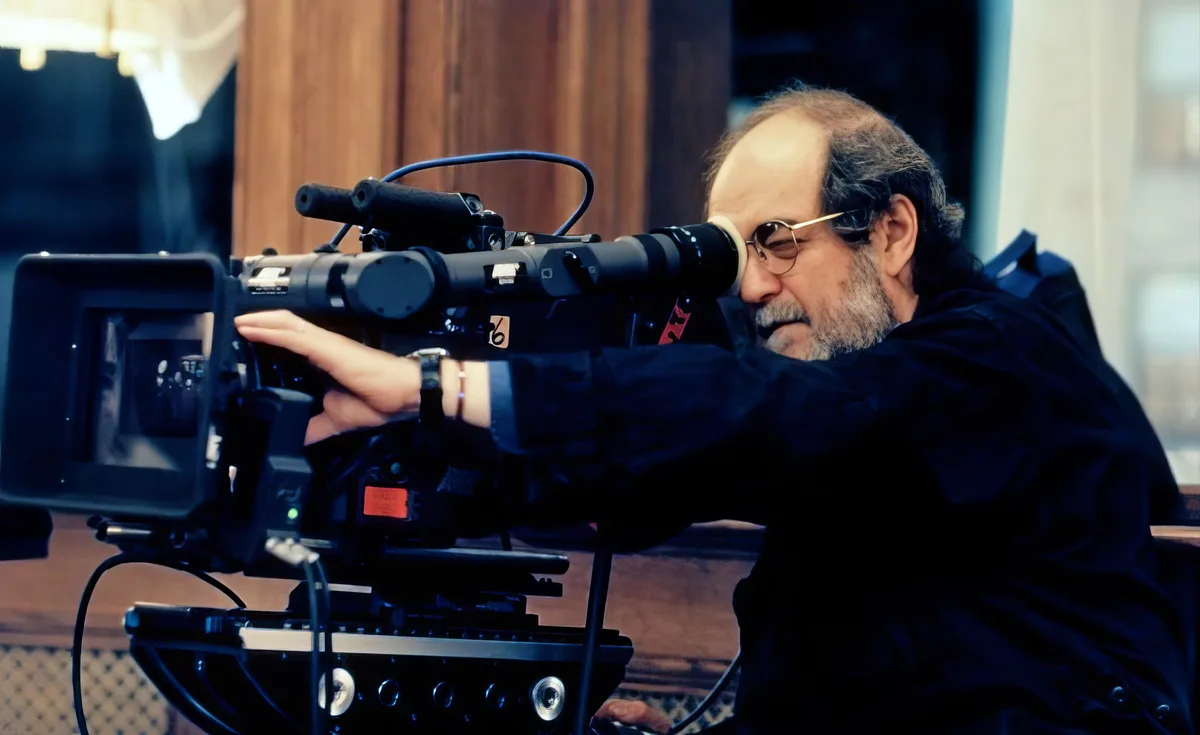 A man with glasses and a beard, wearing a dark shirt, peers through the viewfinder of a large professional film camera. His…