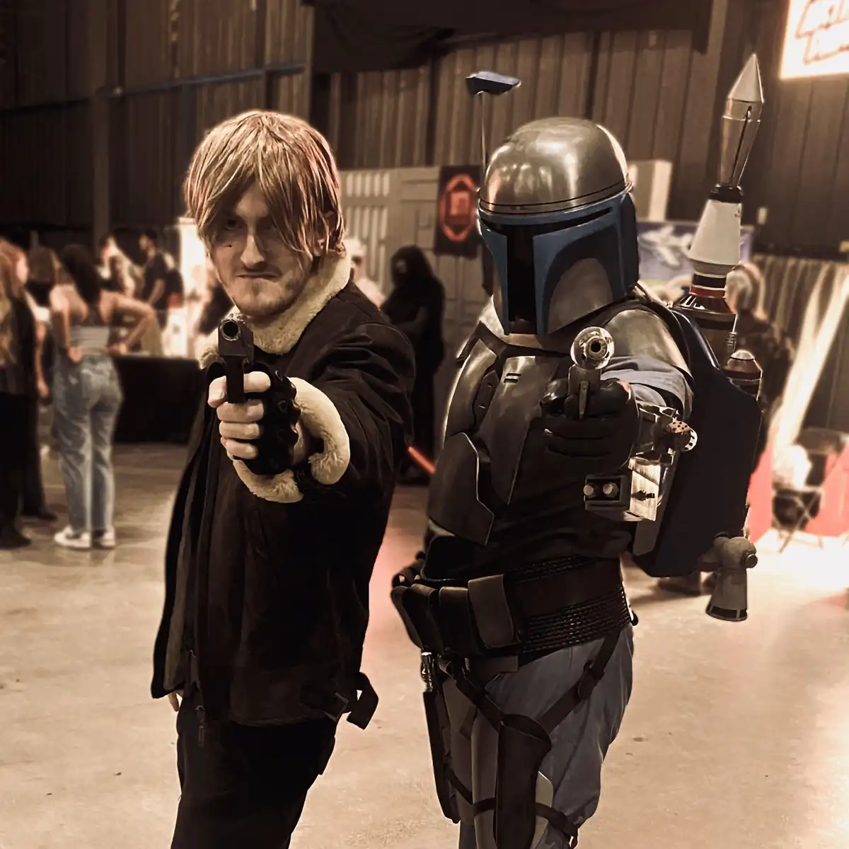 Two people in cosplay stand side-by-side, both pointing handguns directly at the viewer. The person on the left wears a dark…