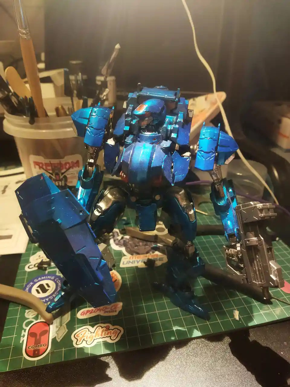 A blue-painted mecha model stands on a green cutting mat surrounded by hobby tools and stickers, with overlaid text reading…
