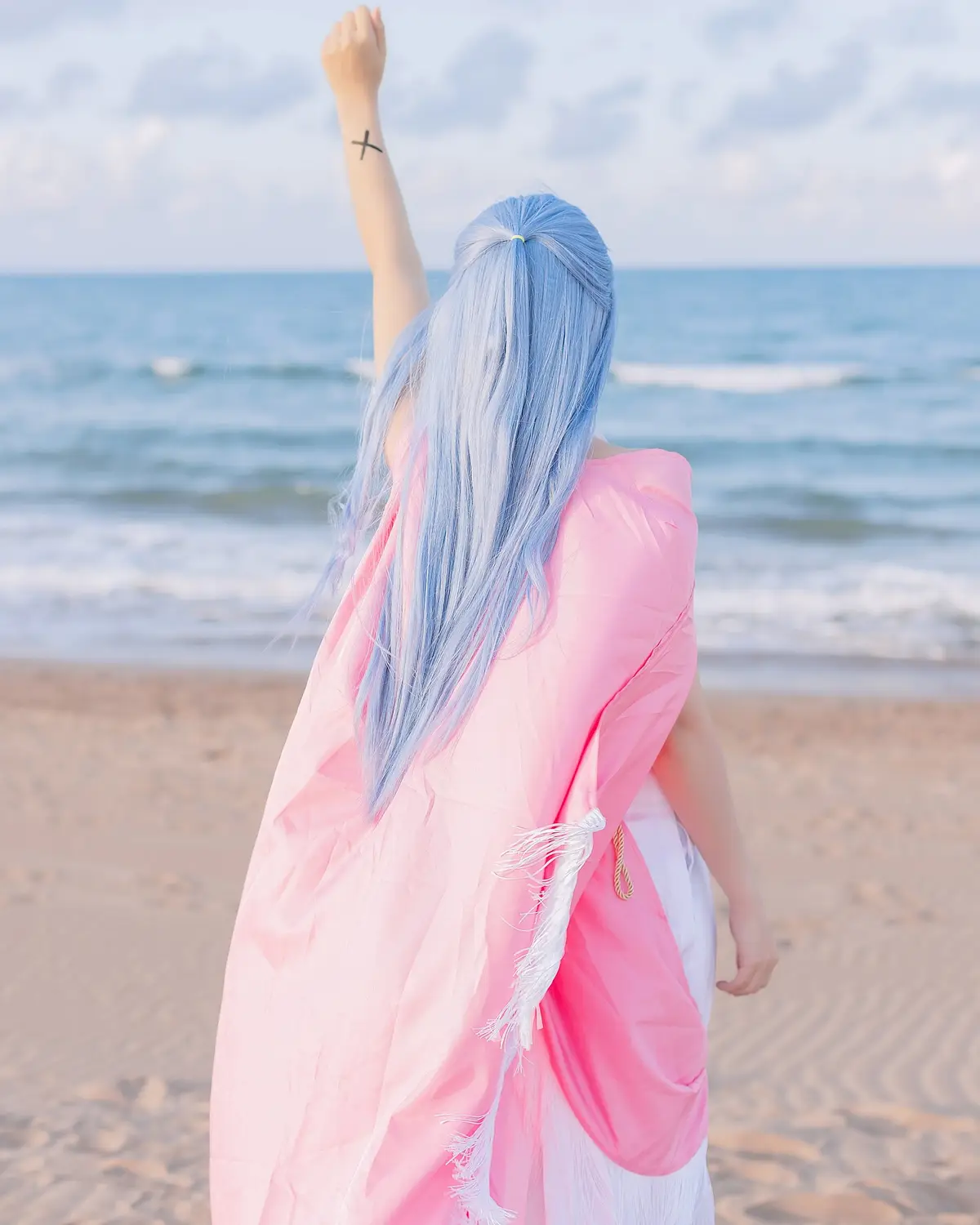 A person with long blue hair, seen from behind, stands on a beach wearing a pink robe over white clothing, raising one arm…
