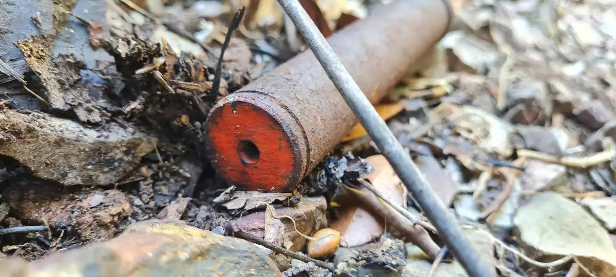 A rusty, cylindrical metal object with a red-painted end and a central hole lies on forest floor littered with dry leaves…