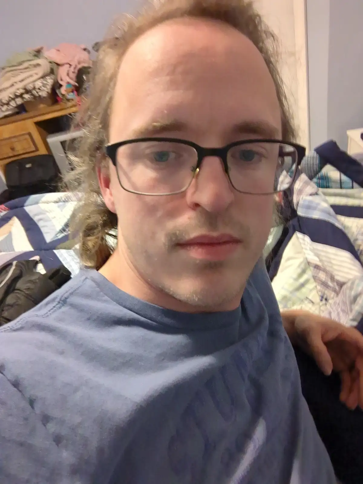 A selfie of a man with glasses and messy hair, wearing a blue t-shirt, looking directly at the camera with a neutral…
