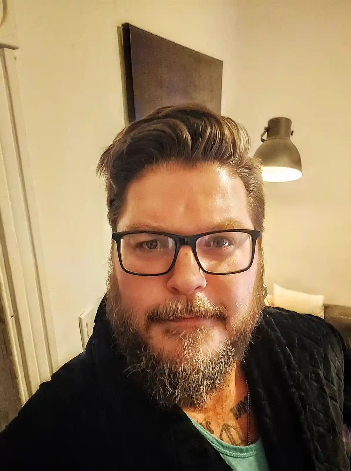 A man with a beard and glasses stares directly at the camera with a neutral expression, wearing a black quilted robe over a…
