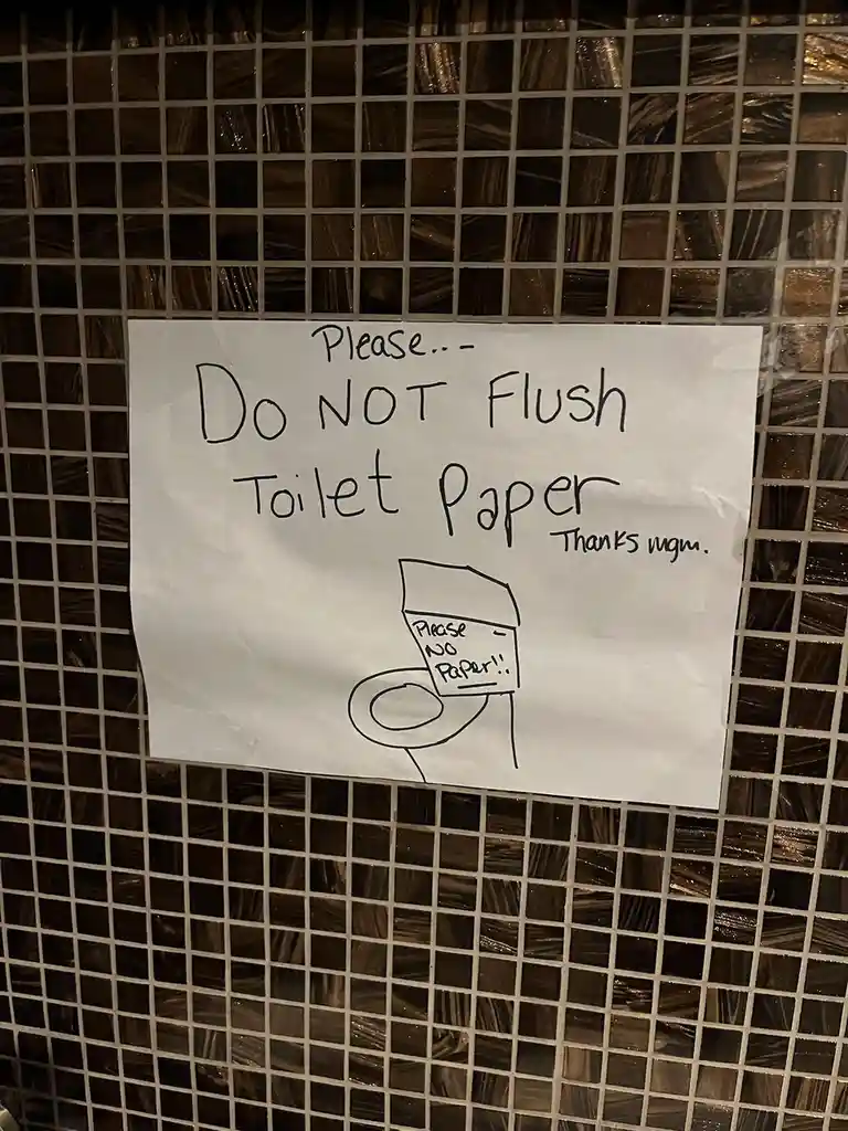 A handwritten sign taped to a dark brown tiled wall in a bathroom, asking people not to flush toilet paper, with a simple…