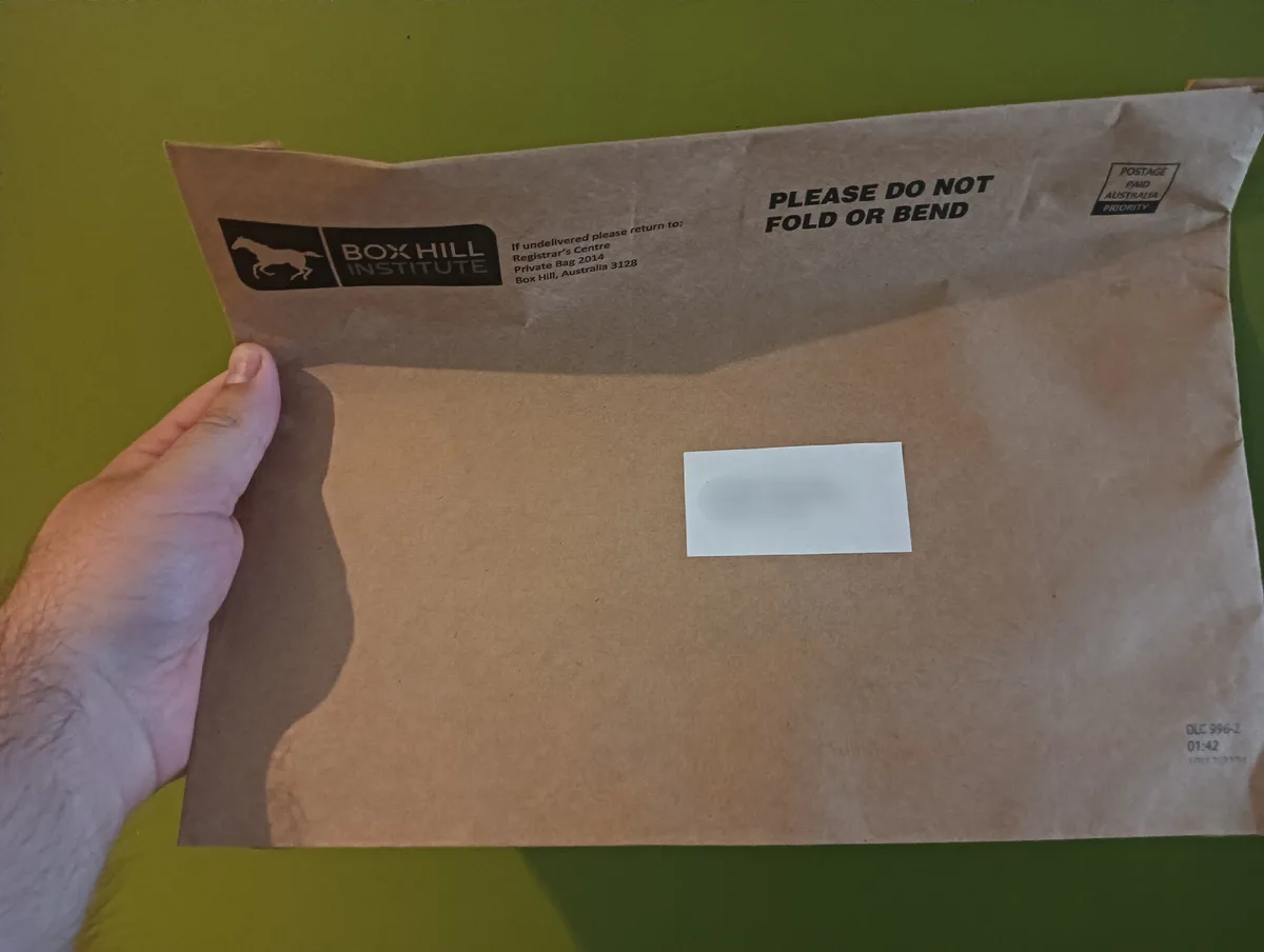 A hand holds a brown paper envelope from Box Hill Institute against a green background. The envelope has a 'PLEASE DO NOT…